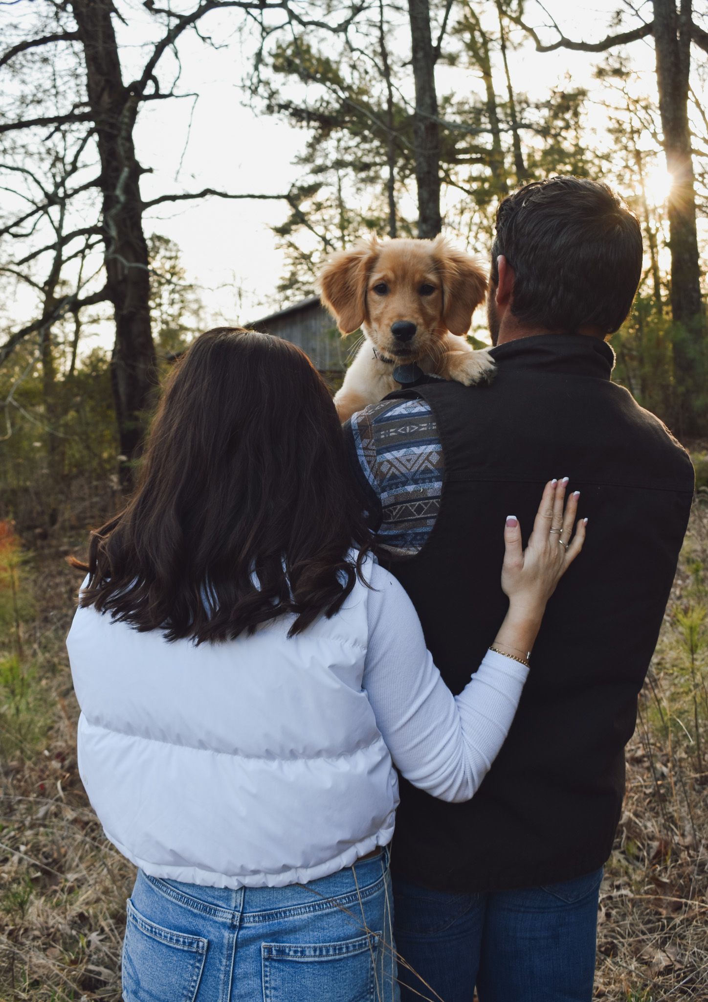 A couple holding their golden retriever puppy while standing in a sunlit forest.