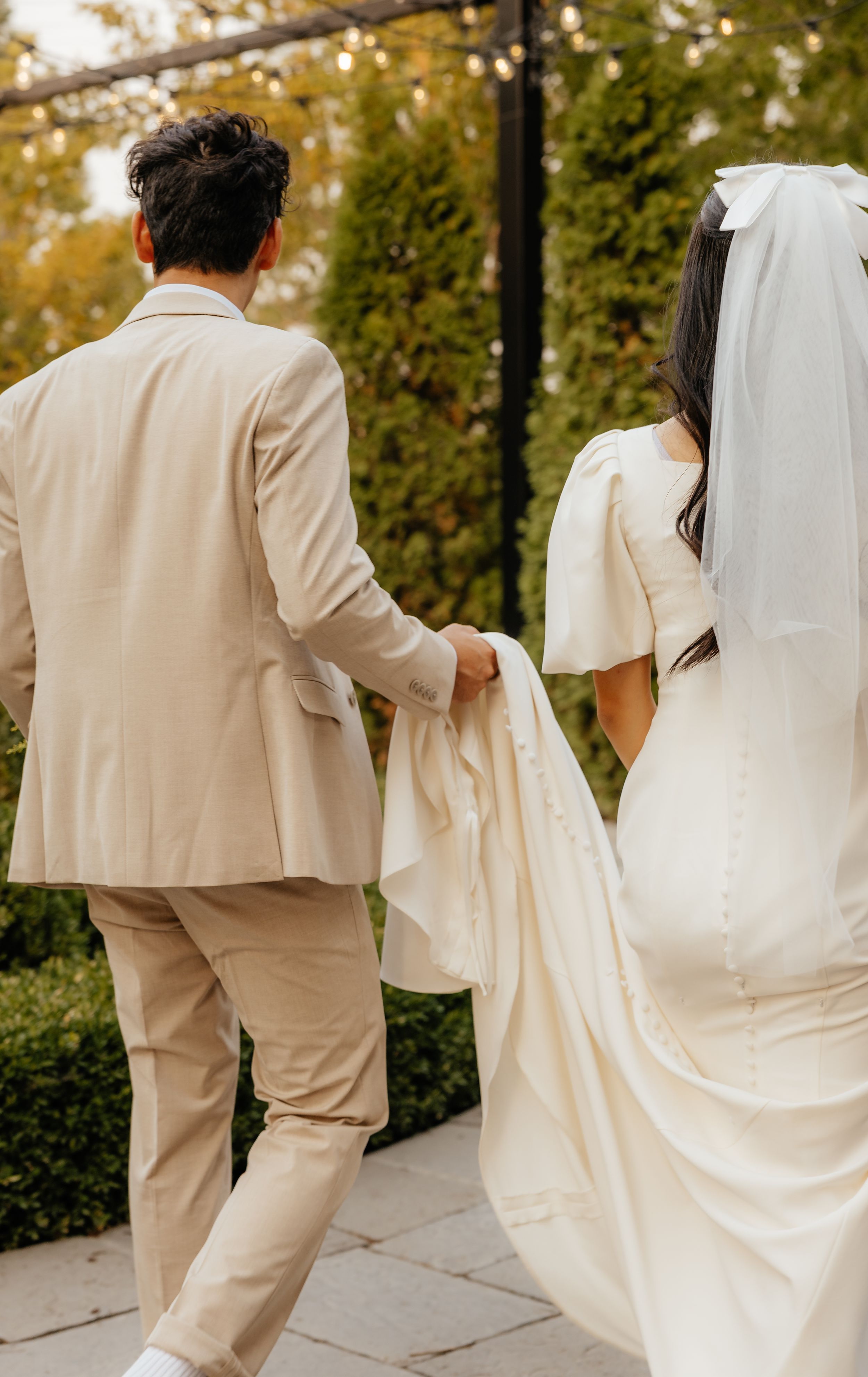 Bride and groom walking hand in hand, groom in beige suit carrying bride's white wedding dress train.