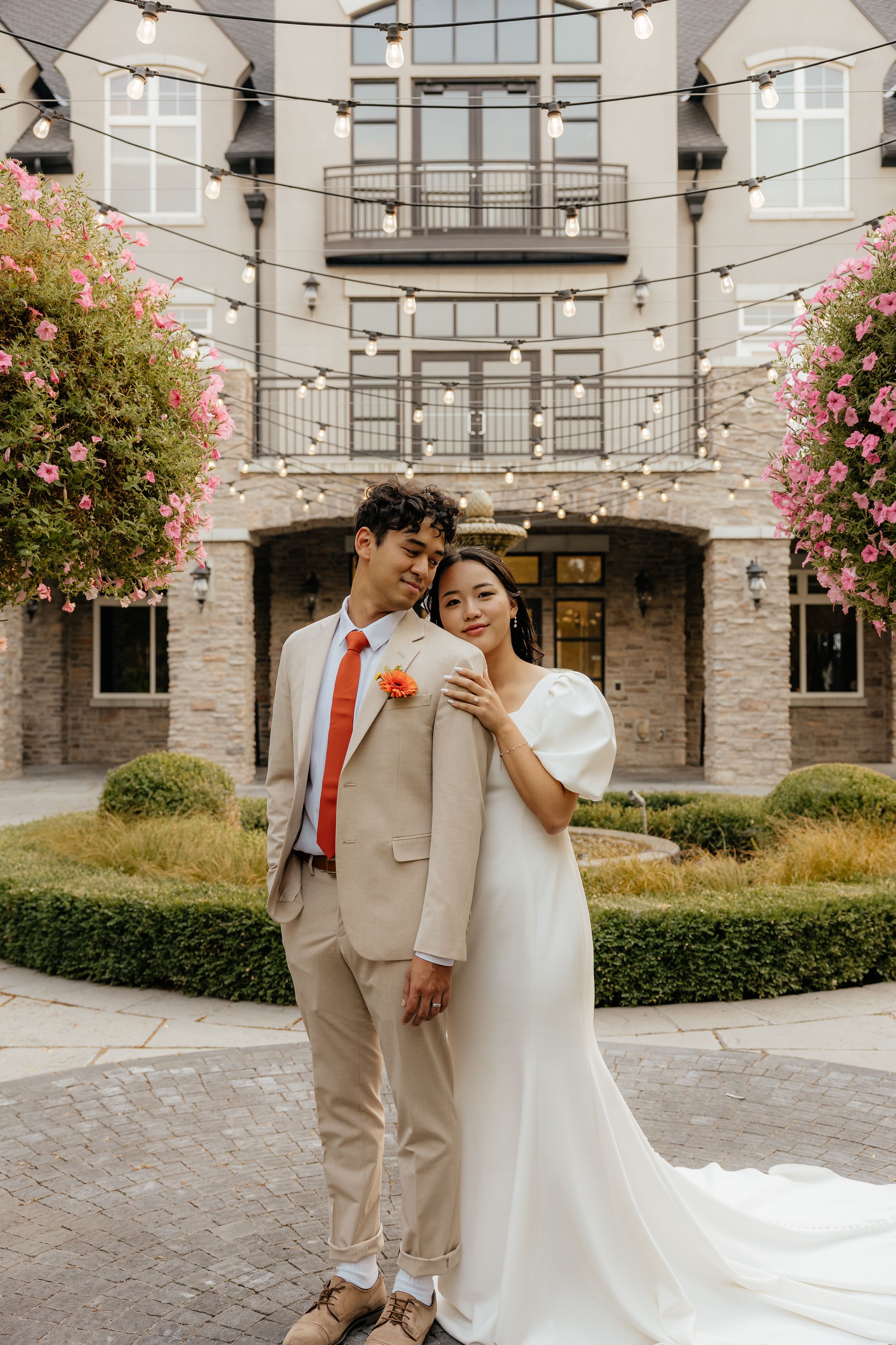 Bride and groom posing in elegant courtyard with string lights and pink flowers, groom in tan suit.