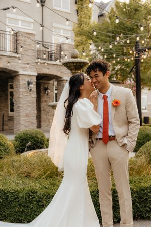 Bride and groom smile together outdoors with string lights and stone building backdrop at elegant wedding venue.