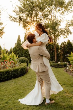 Groom lifts bride in white gown at golden hour in lush garden wedding photo.
