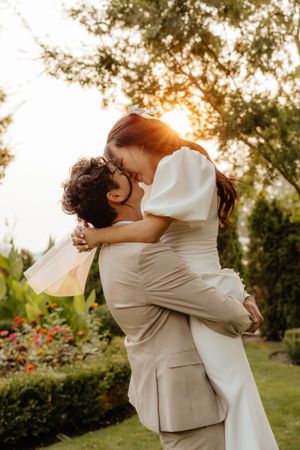 Couple shares romantic kiss as groom lifts bride in white dress during golden sunset wedding.