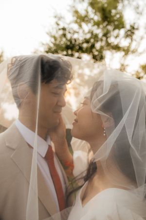 Bride and groom share intimate moment under flowing veil with warm sunset light.