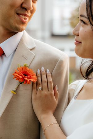 Close-up of bride\u2019s engagement ring as she rests hand on groom\u2019s tan suit with orange boutonniere.