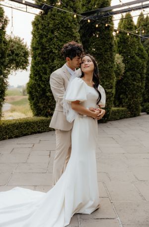 Groom embraces bride from behind under pergola with tall hedges at outdoor wedding venue.