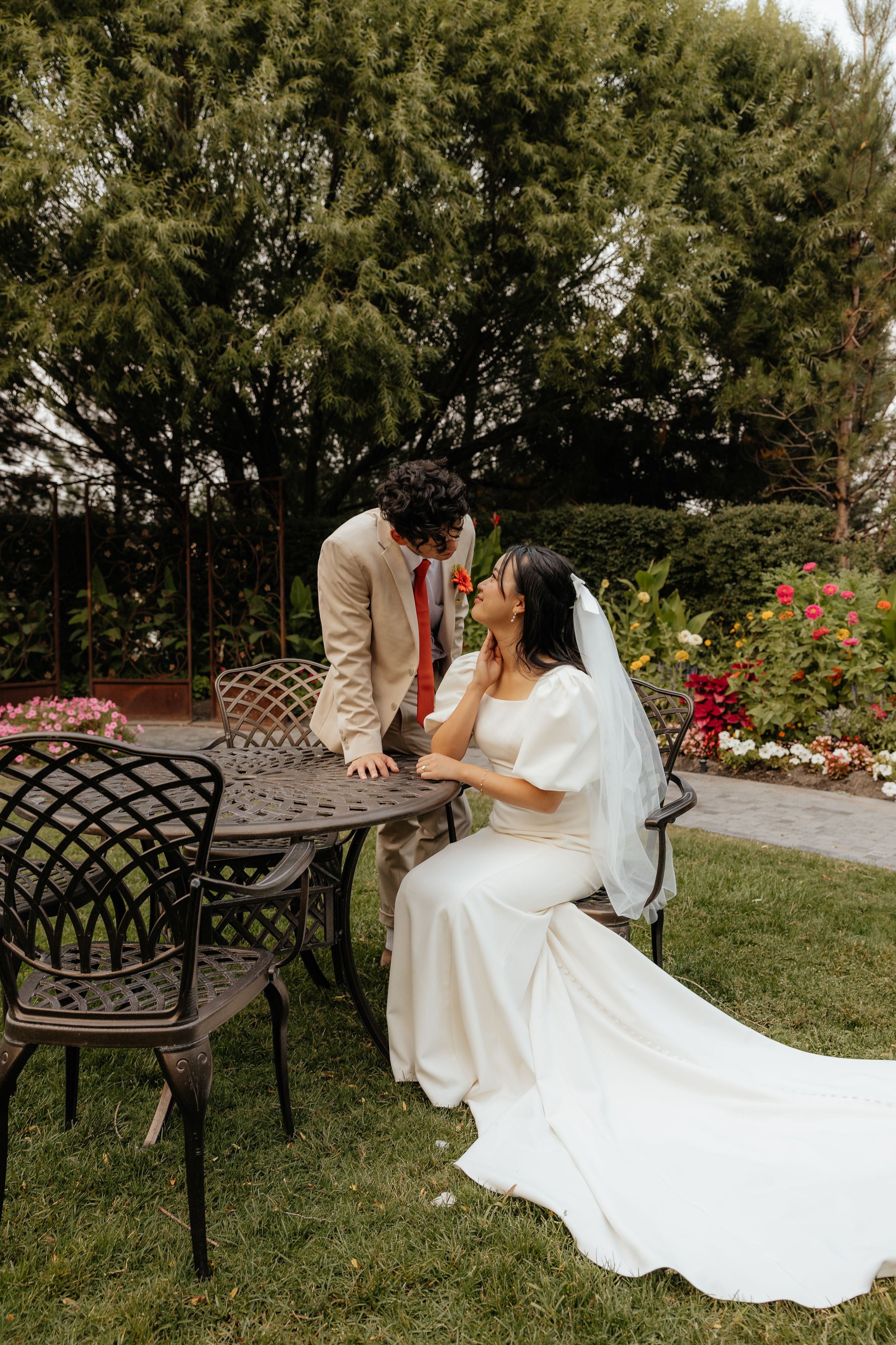 Bride sits at ornate garden table smiling up at groom leaning in for a kiss.