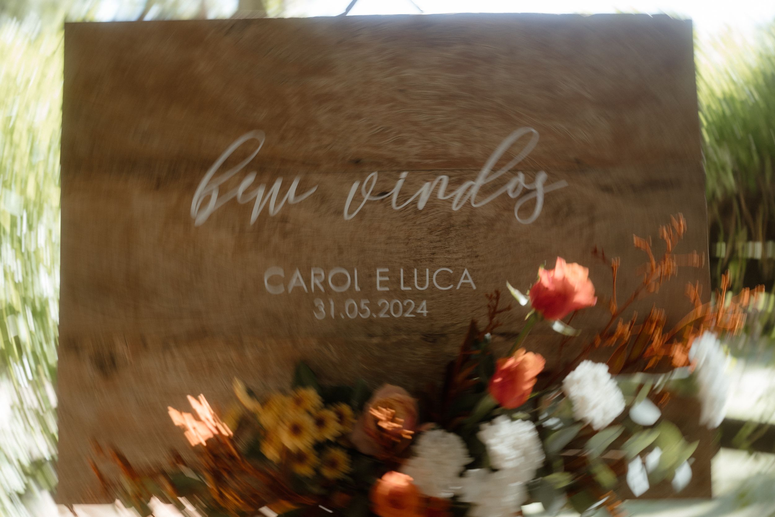 Rustic wooden wedding sign with floral decorations displaying 'Carol e Luca' and a date.