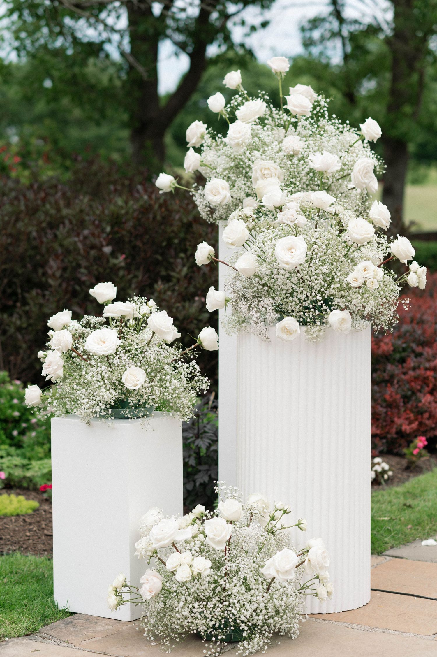 Elegant white floral arrangements with roses and baby's breath on white pedestals in garden setting.