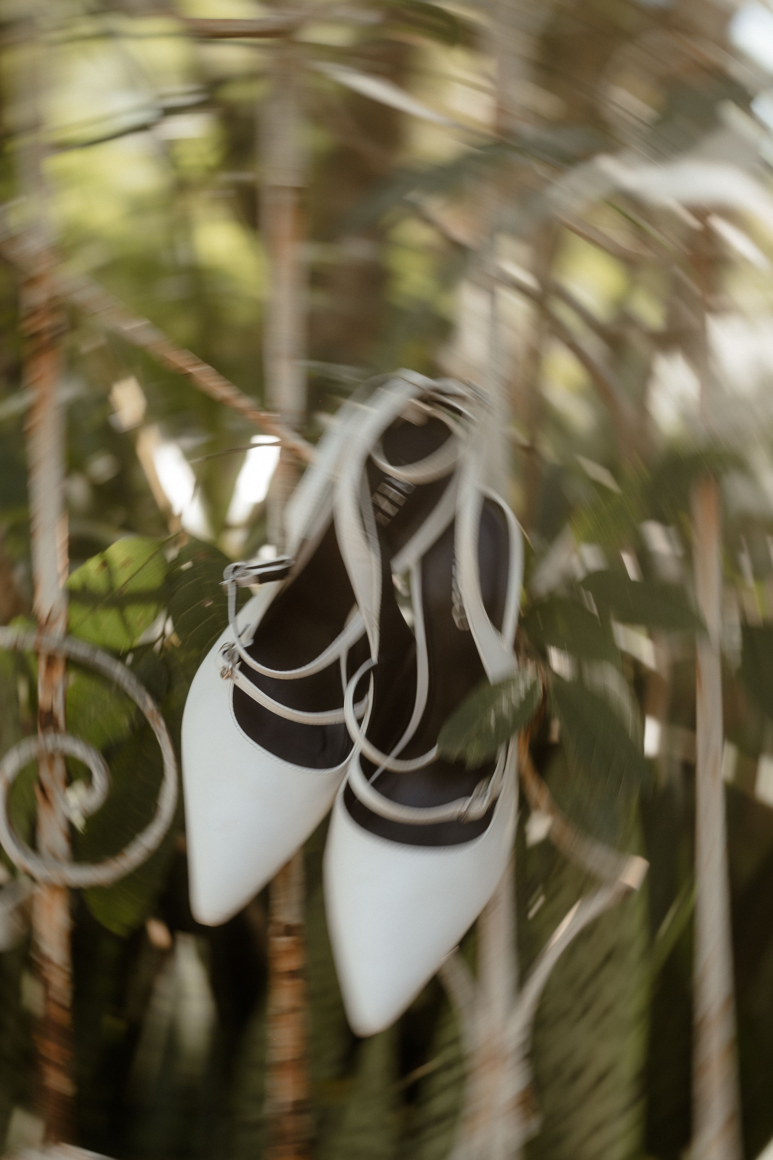 White bridal shoes with ribbon ties hanging against blurred botanical background.