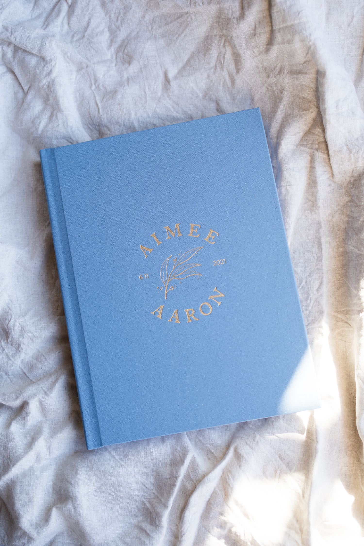 Light blue wedding album with gold lettering on wrinkled white fabric.