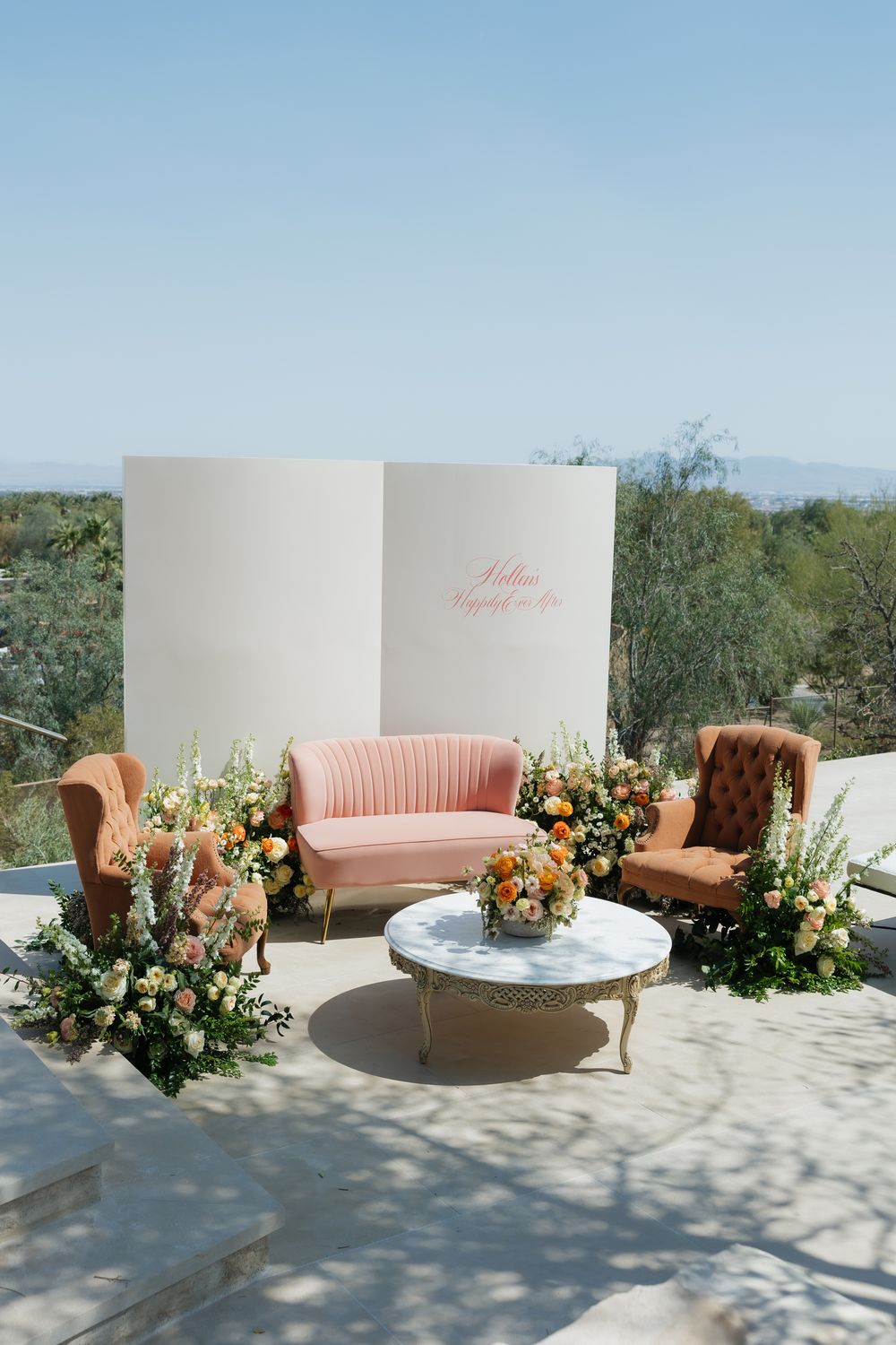 Elegant outdoor lounge setup with pink velvet sofa, vintage chairs, and floral arrangements against white backdrop.