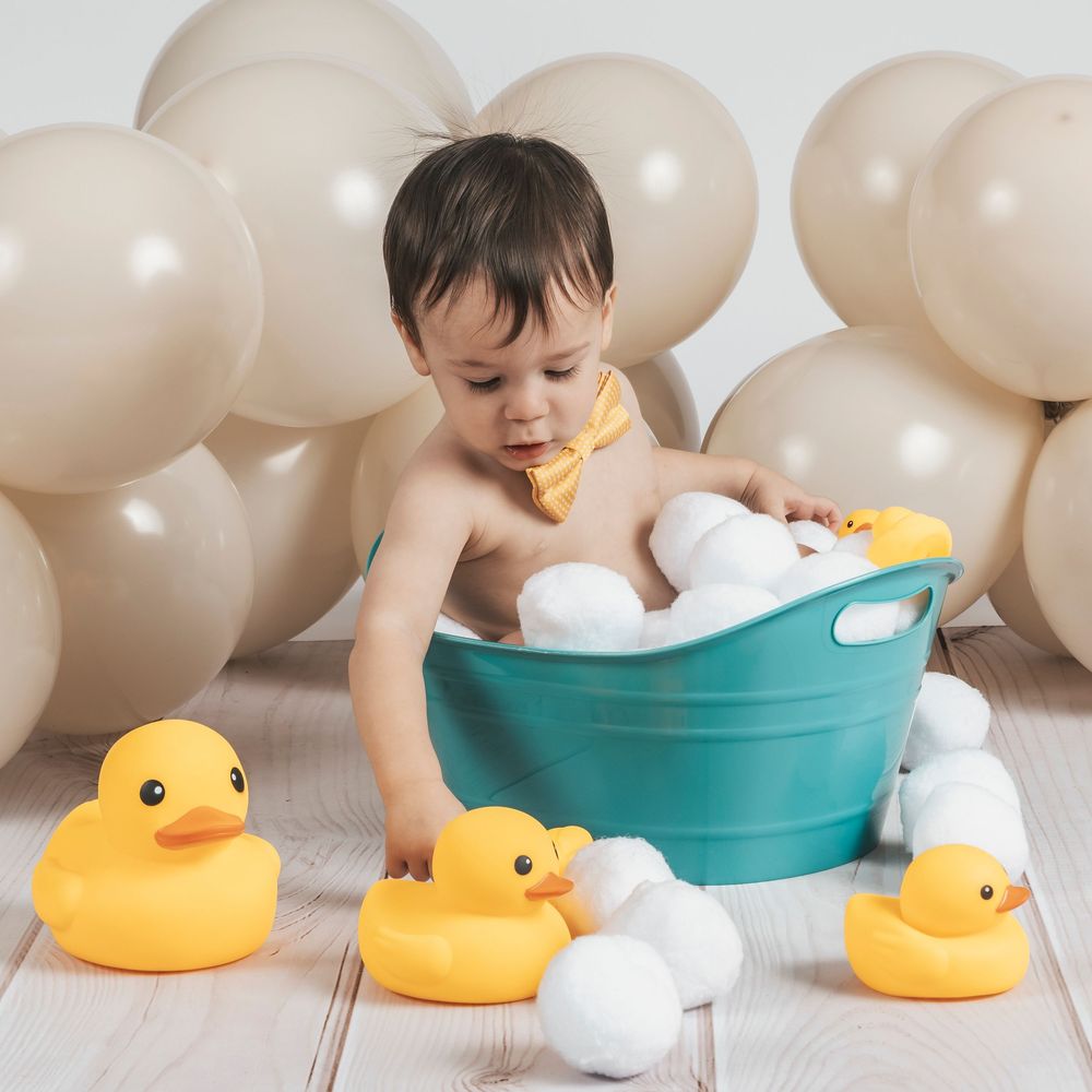 Baby boy plays with yellow rubber ducks beside a teal tub filled with bubbles and balloons.