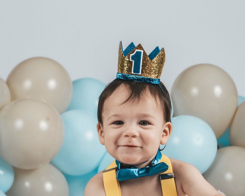 Baby wearing gold glitter crown and suspenders celebrating first birthday with balloons.