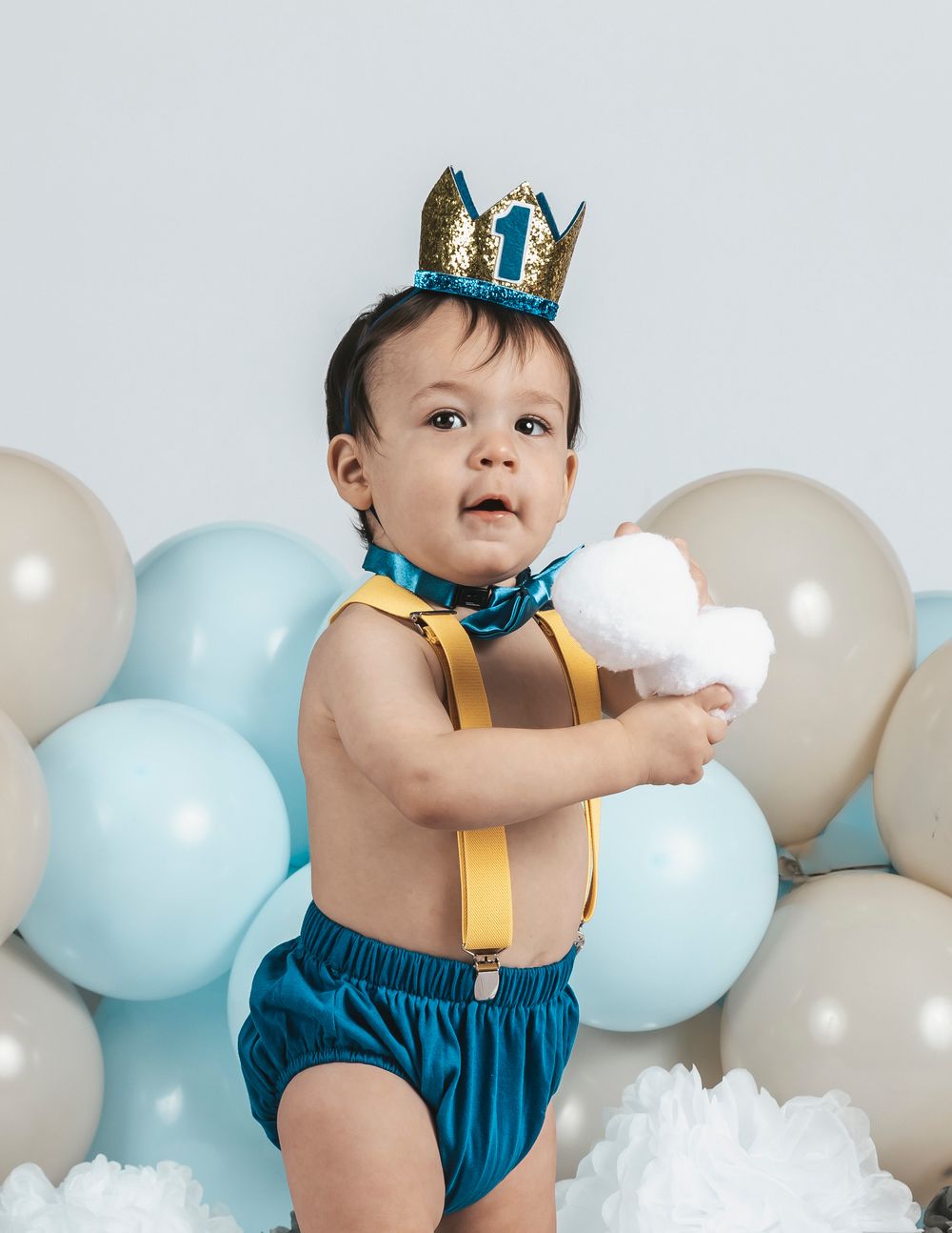 Baby boy in teal bloomers and gold crown celebrates first birthday surrounded by pastel balloons.