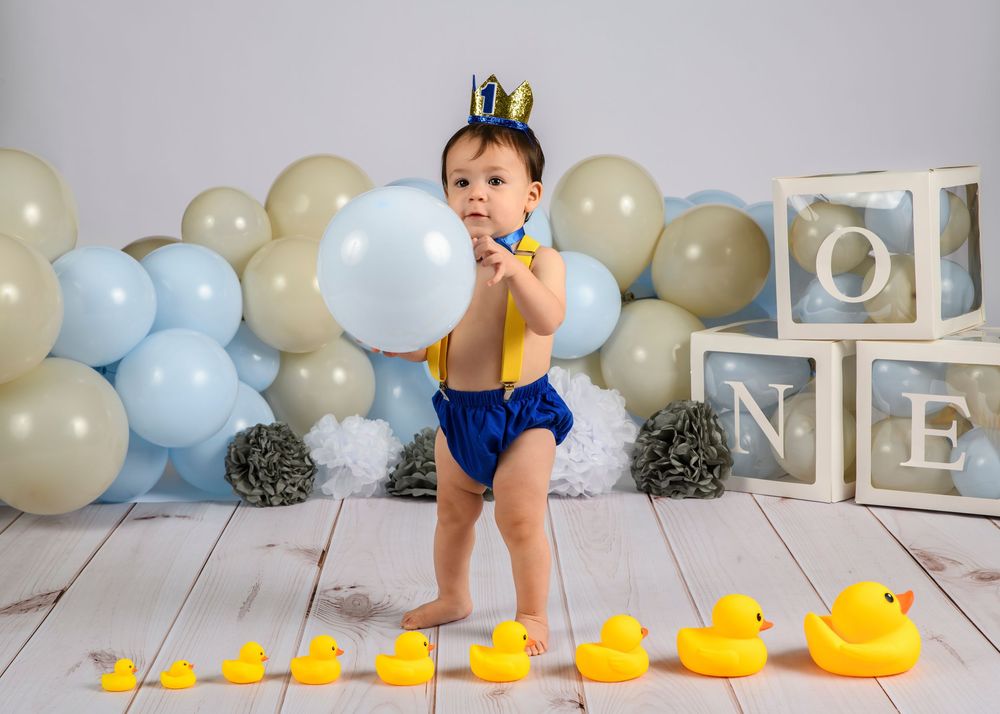 One-year-old boy in suspenders holds balloon near rubber ducks and ONE blocks at birthday photoshoot.
