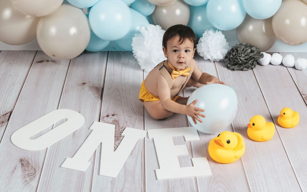 Baby in yellow onesie with angel wings sits beside ONE letters and rubber ducks with balloons.
