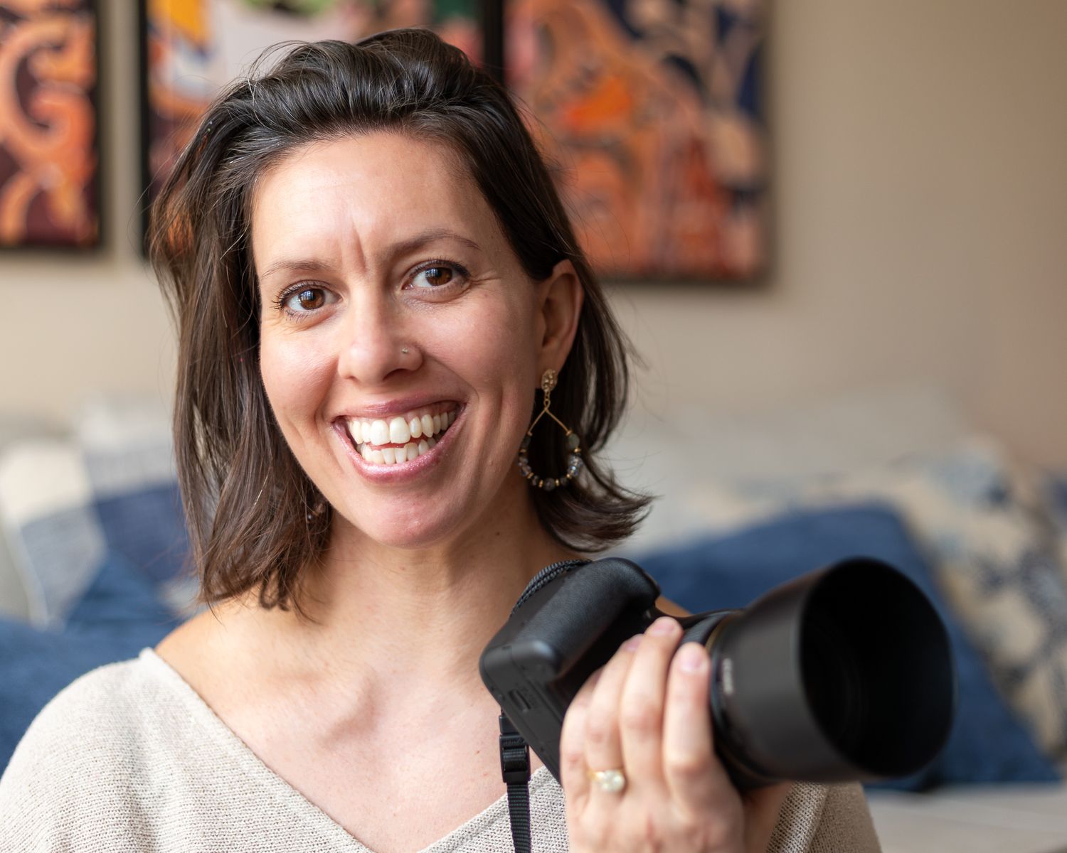 A woman with a large open smile holding her camera posing against a blurred background.