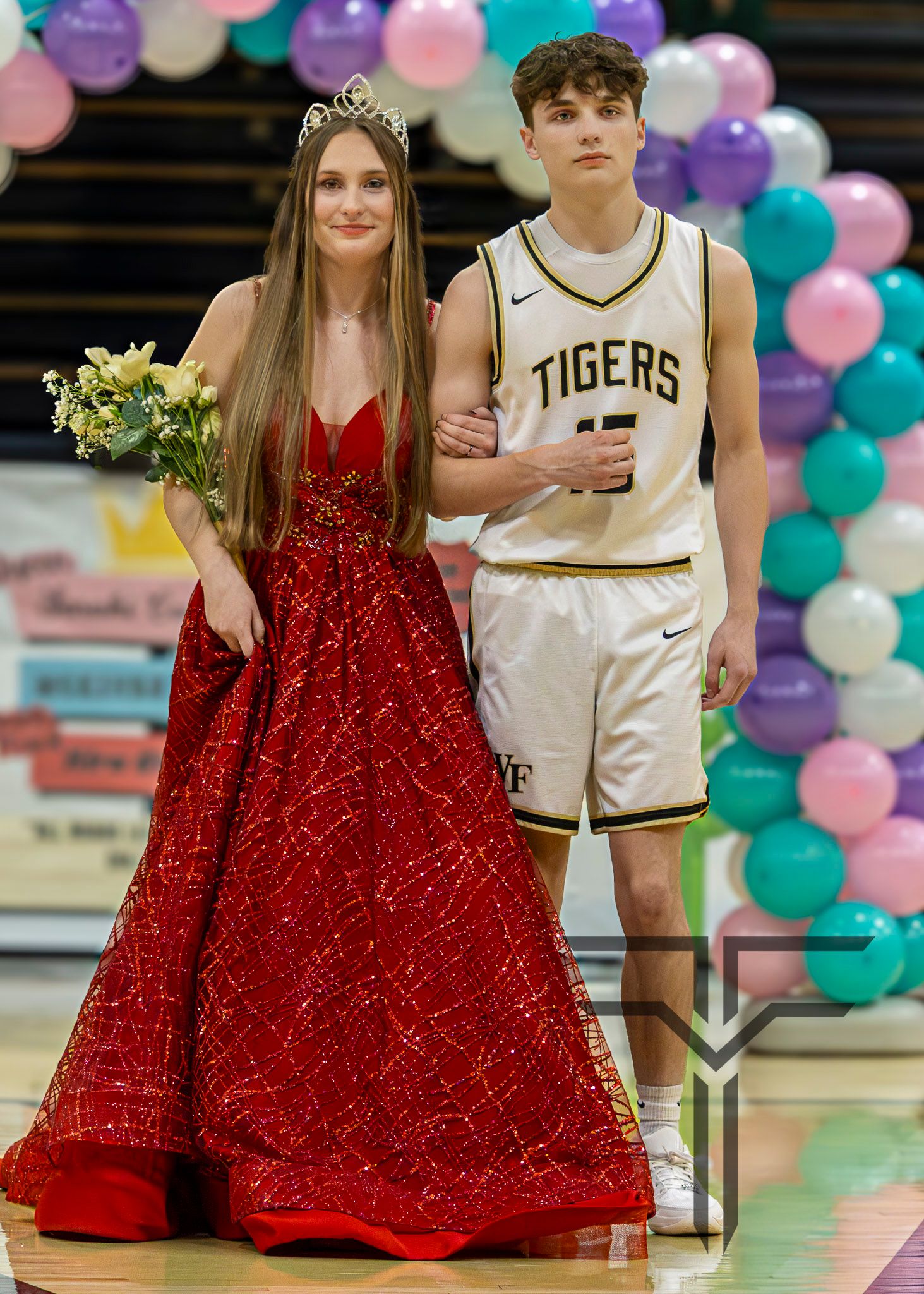 Homecoming queen in red glitter gown with crown and bouquet stands with Tigers basketball player at school gym ceremony.