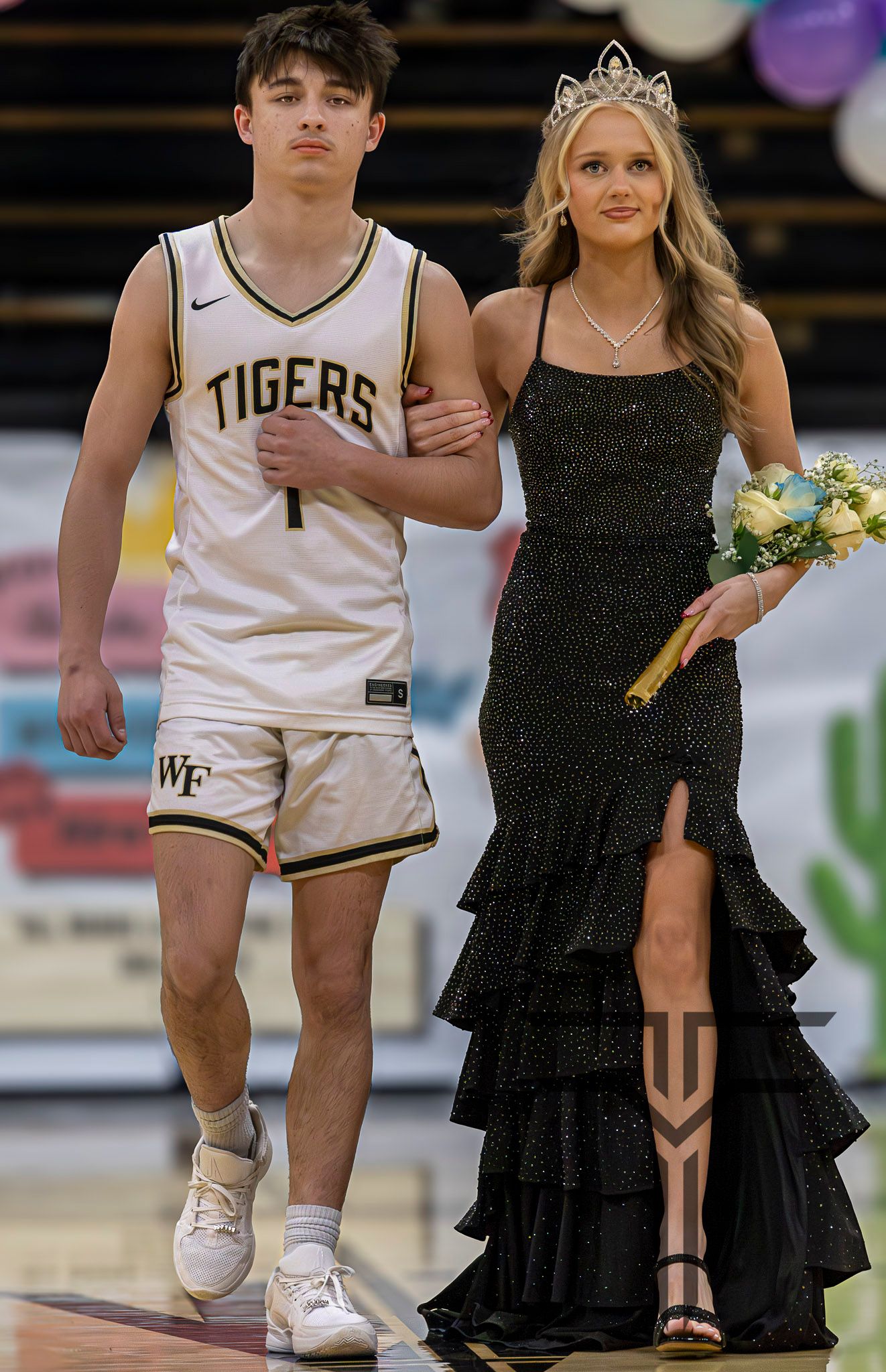A teenage boy in a Tigers basketball uniform escorts a girl wearing a black sparkly gown and tiara at a homecoming ceremony.