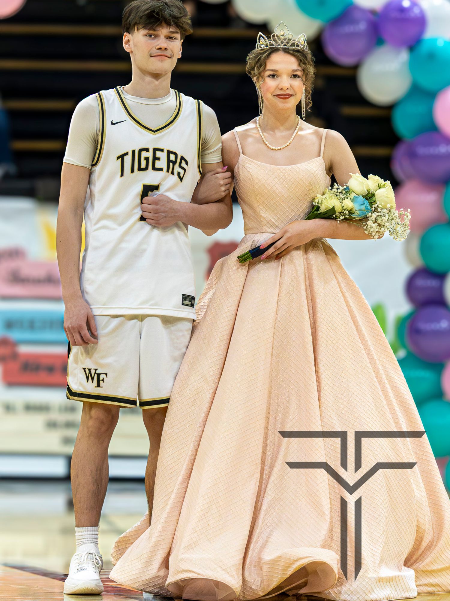 A young couple poses at a school event, he in a Tigers basketball uniform, she in a elegant peach ball gown with a tiara.