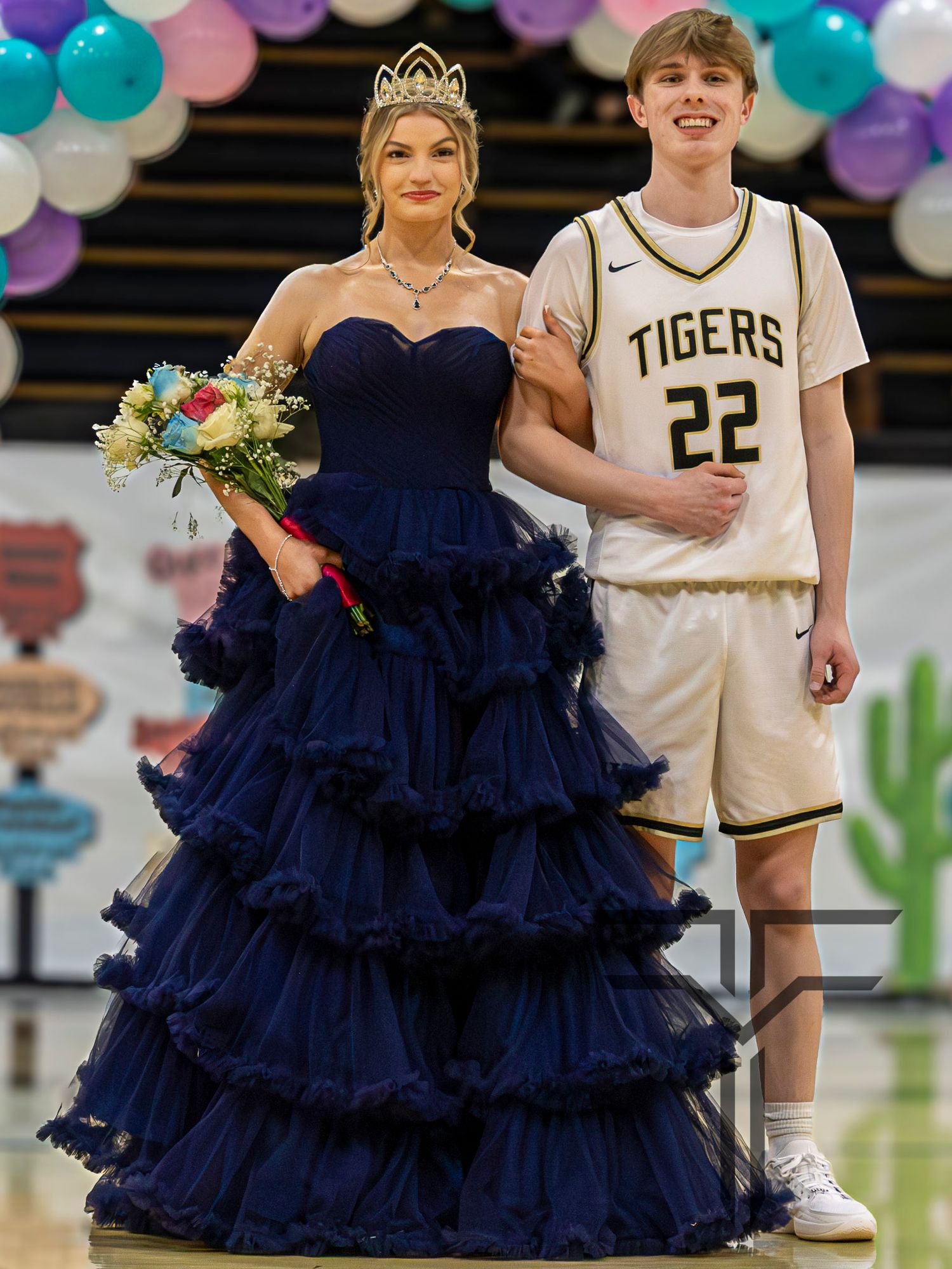 Blonde girl in navy blue ruffled ball gown and crown holds bouquet with Tigers player number 22 at homecoming event.