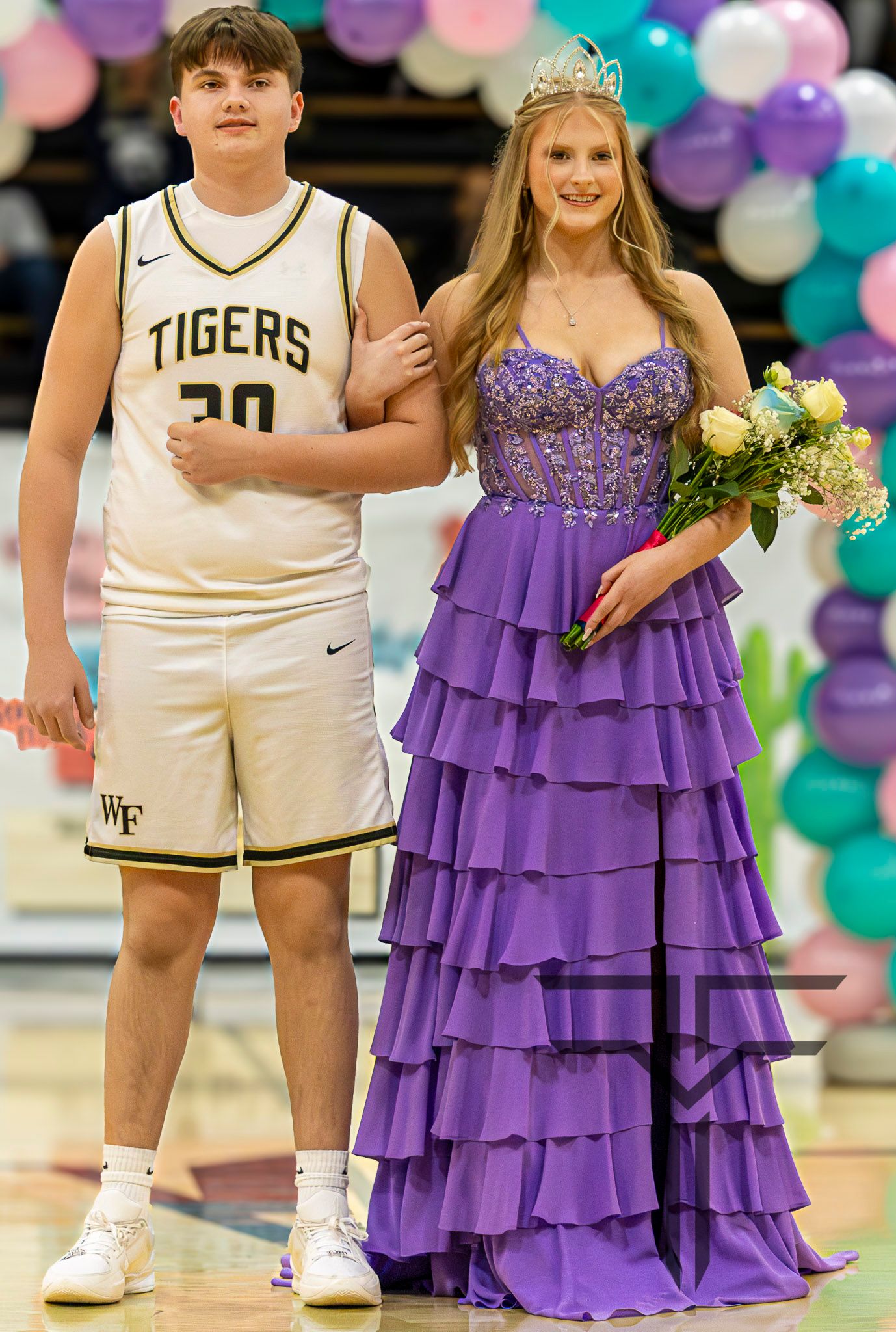 A young man in a Tigers basketball jersey escorts a girl in a purple ruffled gown wearing a crown at a school event.