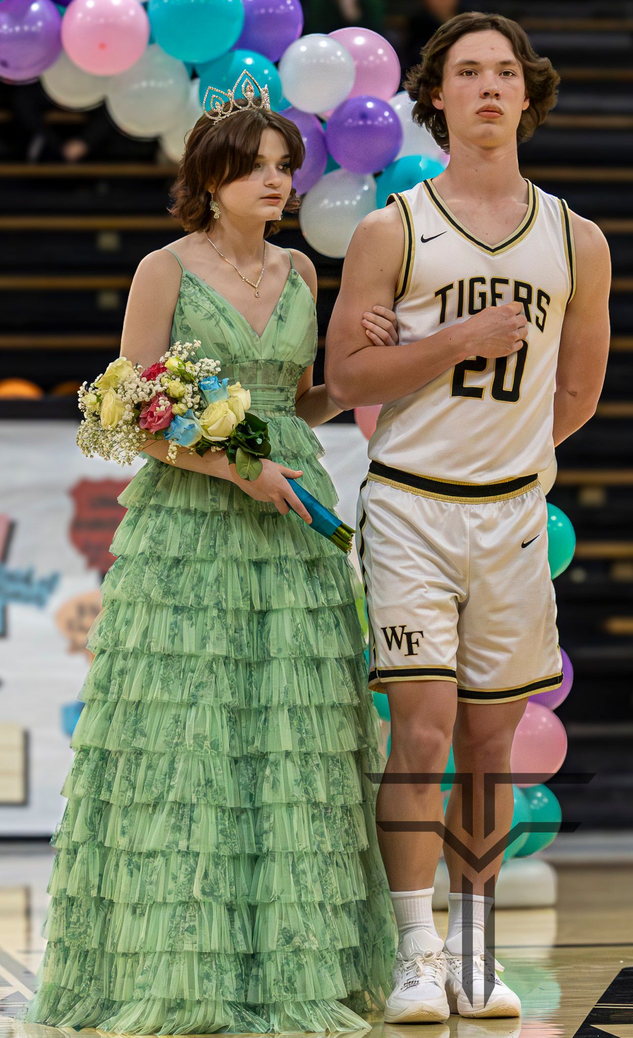Young woman in green ruffled gown and tiara holds bouquet alongside Tigers basketball player number 20 at homecoming ceremony.