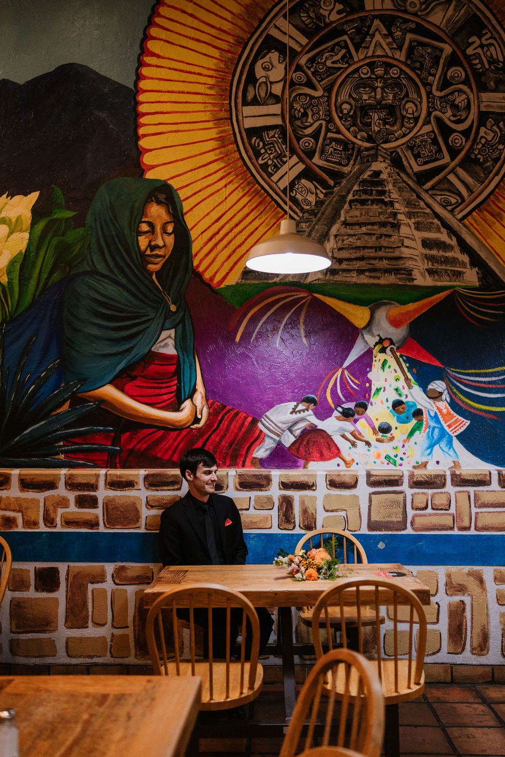 Mexican restaurant interior with colorful mural featuring Aztec calendar and vibrant artwork above stone wall and wooden tables.