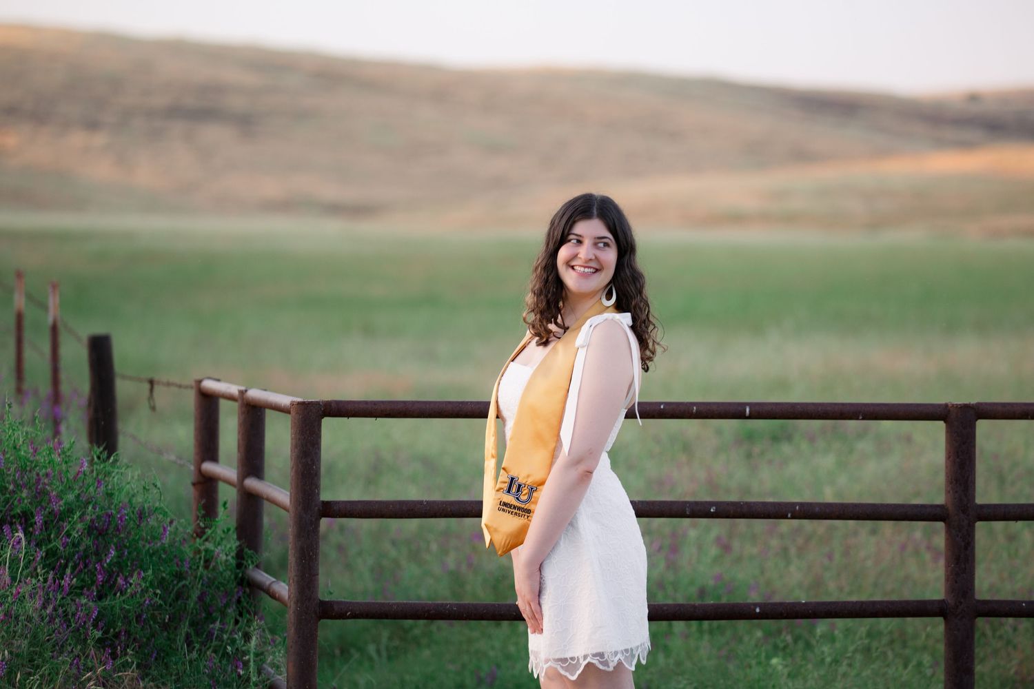 A couple in casual attire stands together in a grassy field with wooden fencing at sunset.