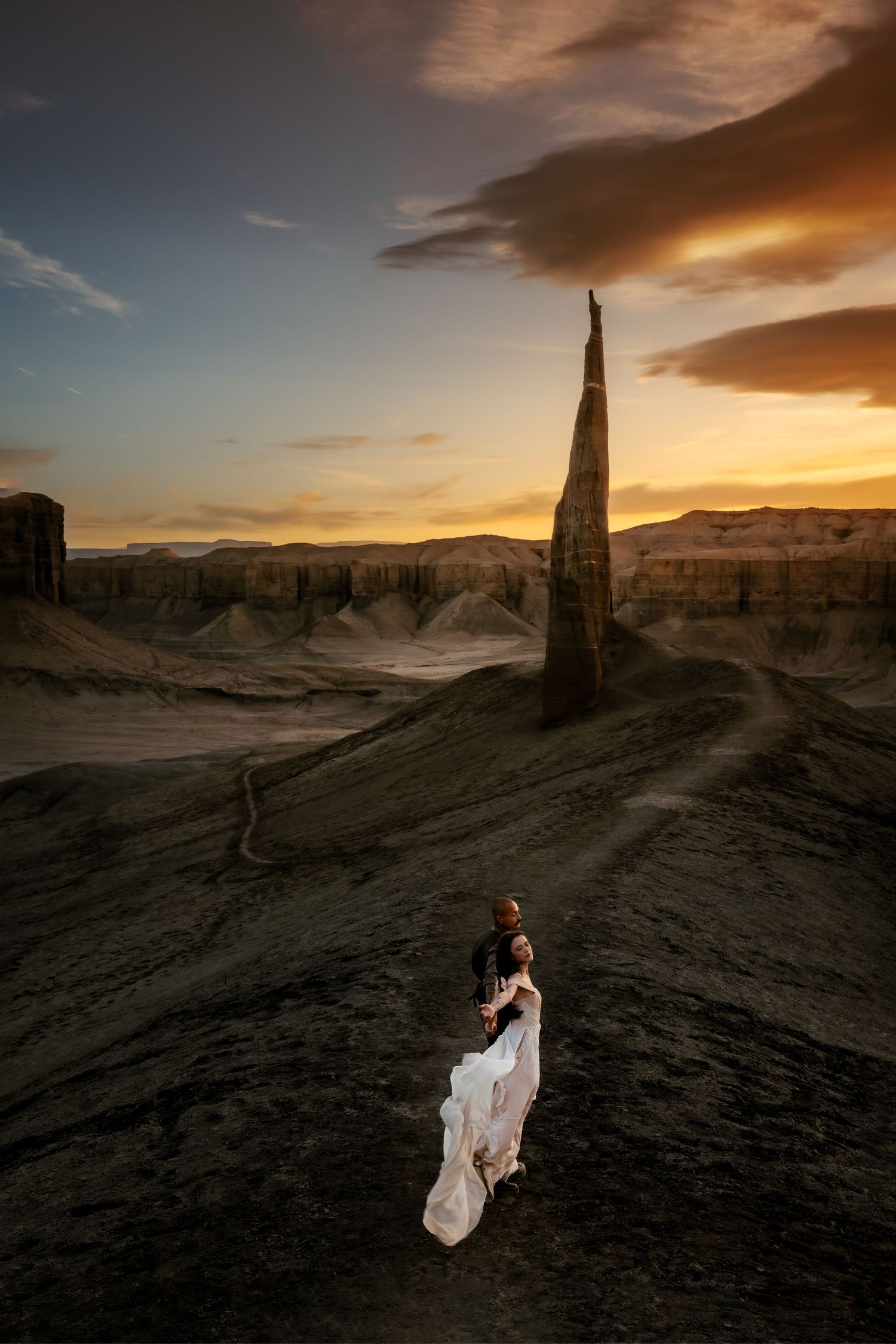 Dramatic desert landscape at sunset with towering rock formations and a figure in flowing white dress creating ethereal silhouettes.