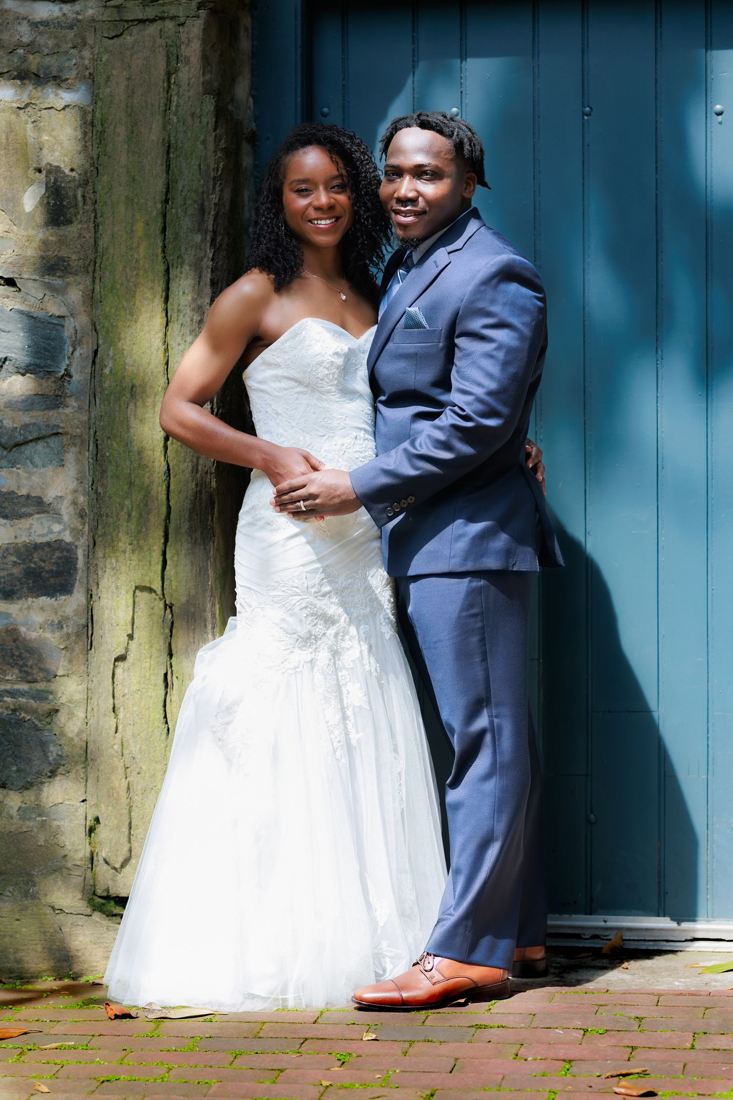 A couple in wedding attire sits on brick steps, with the bride in white dress and groom in navy suit.