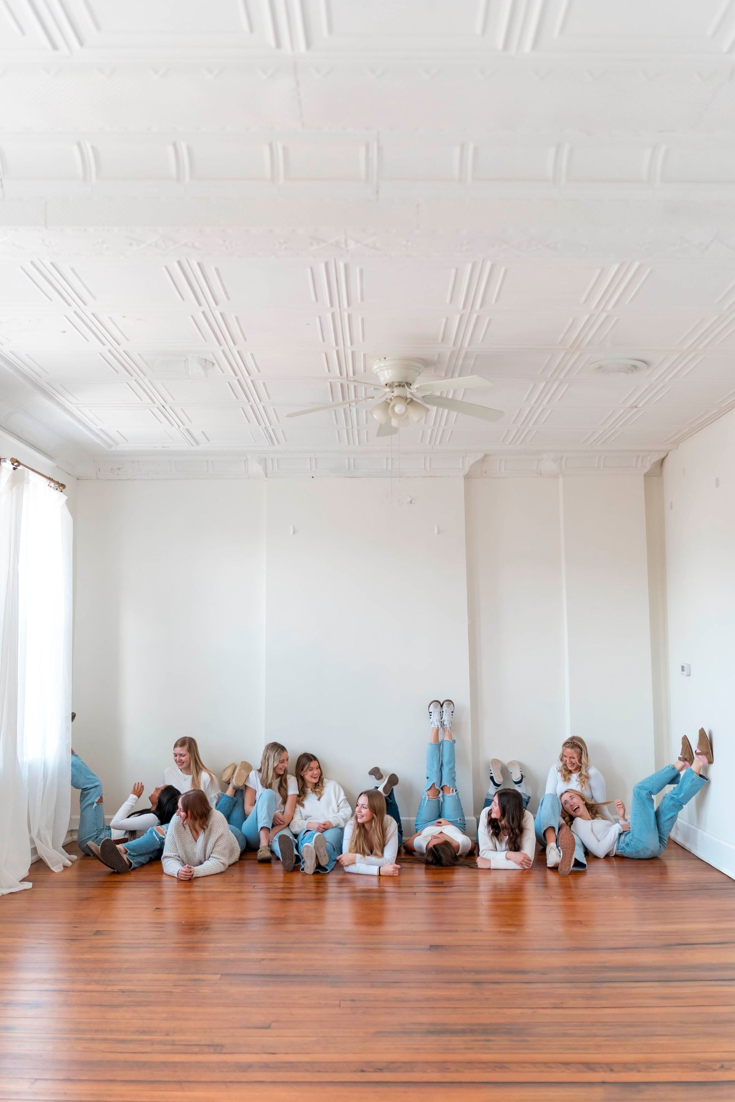 People in light blue clothing lie along a wall in a bright white studio with wooden floors in an artistic arrangement.
