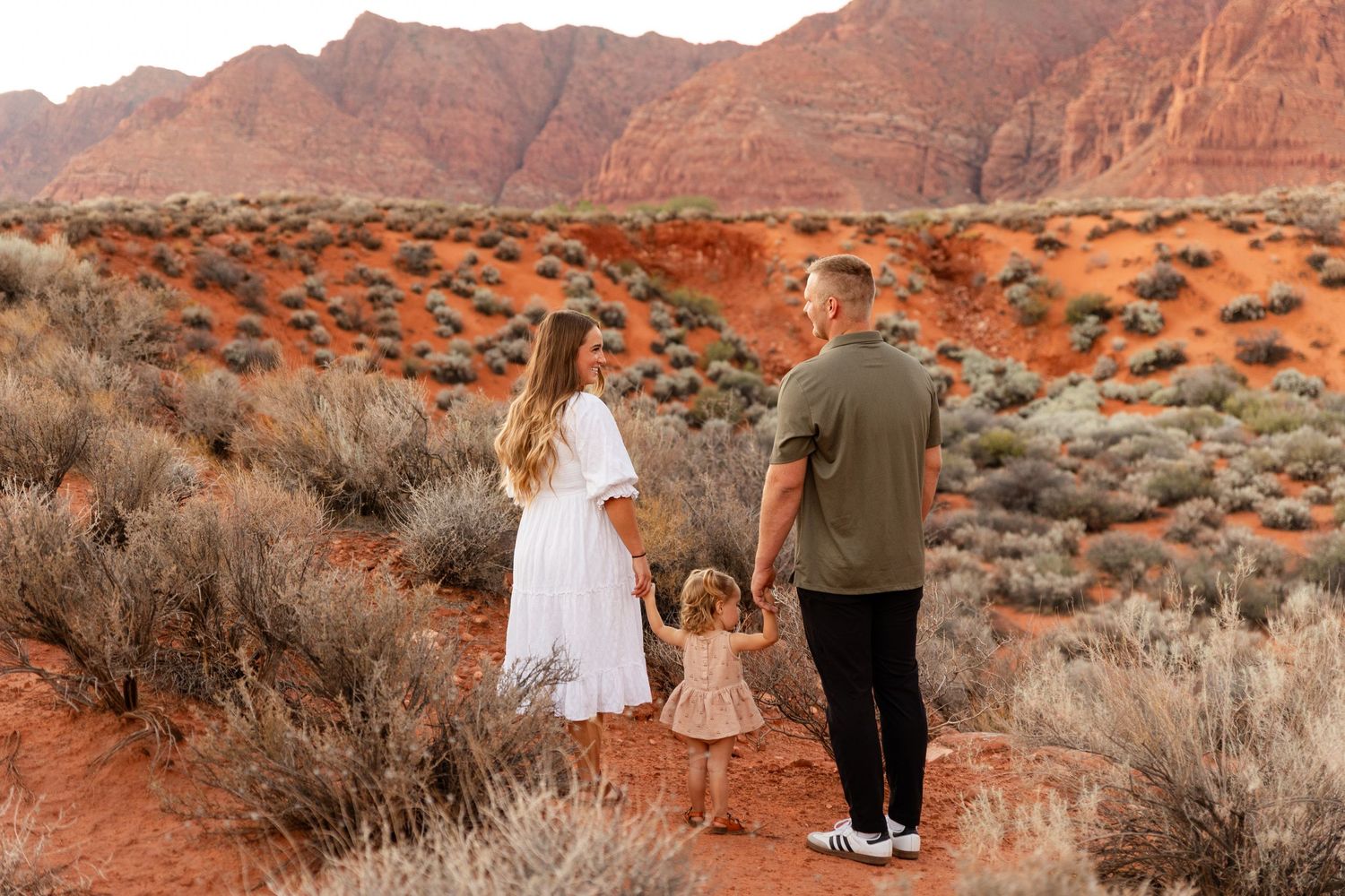 mom and dad holding little girls hand as if walking away towards a beautiful red mountain.