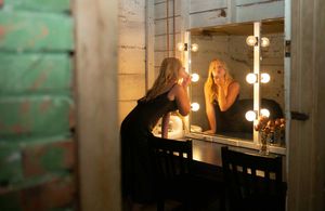 A person sits at a vanity mirror with glowing lights in a dimly lit dressing room.