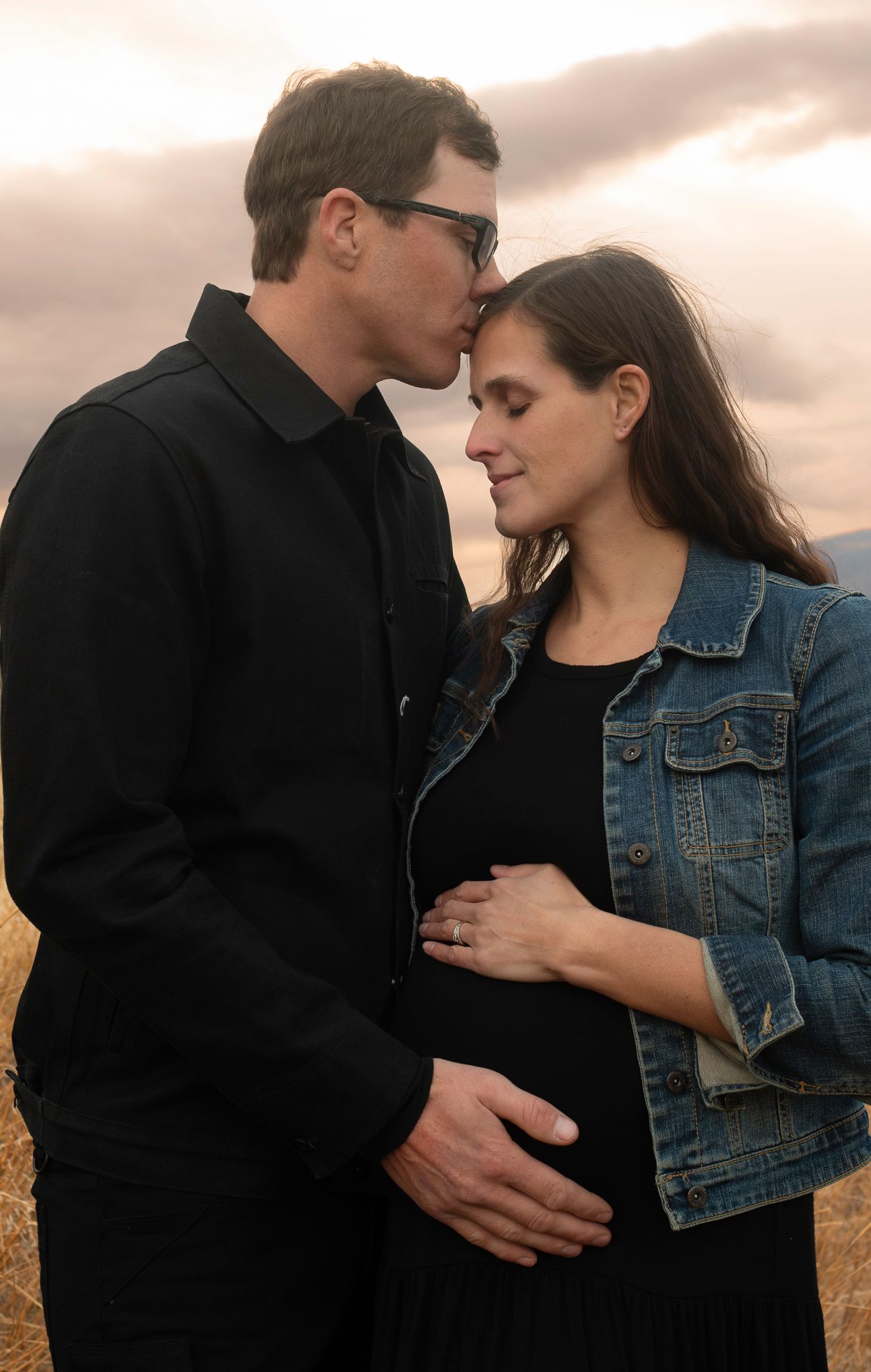 Romantic couple embracing in a field at sunset wearing denim jacket and black clothing.