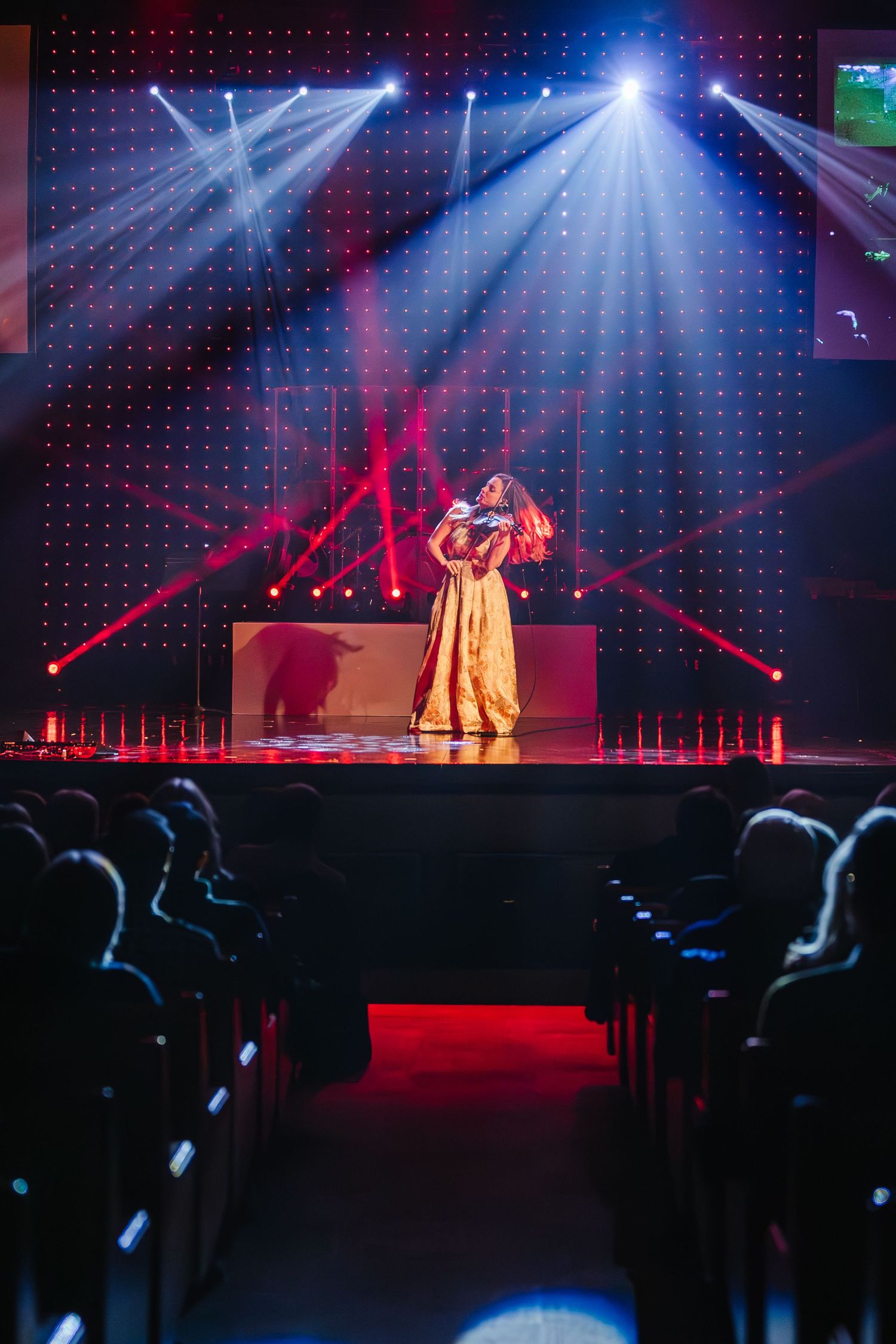 Stage performers dance under dramatic blue and red lighting at a concert venue with silhouetted audience members visible.