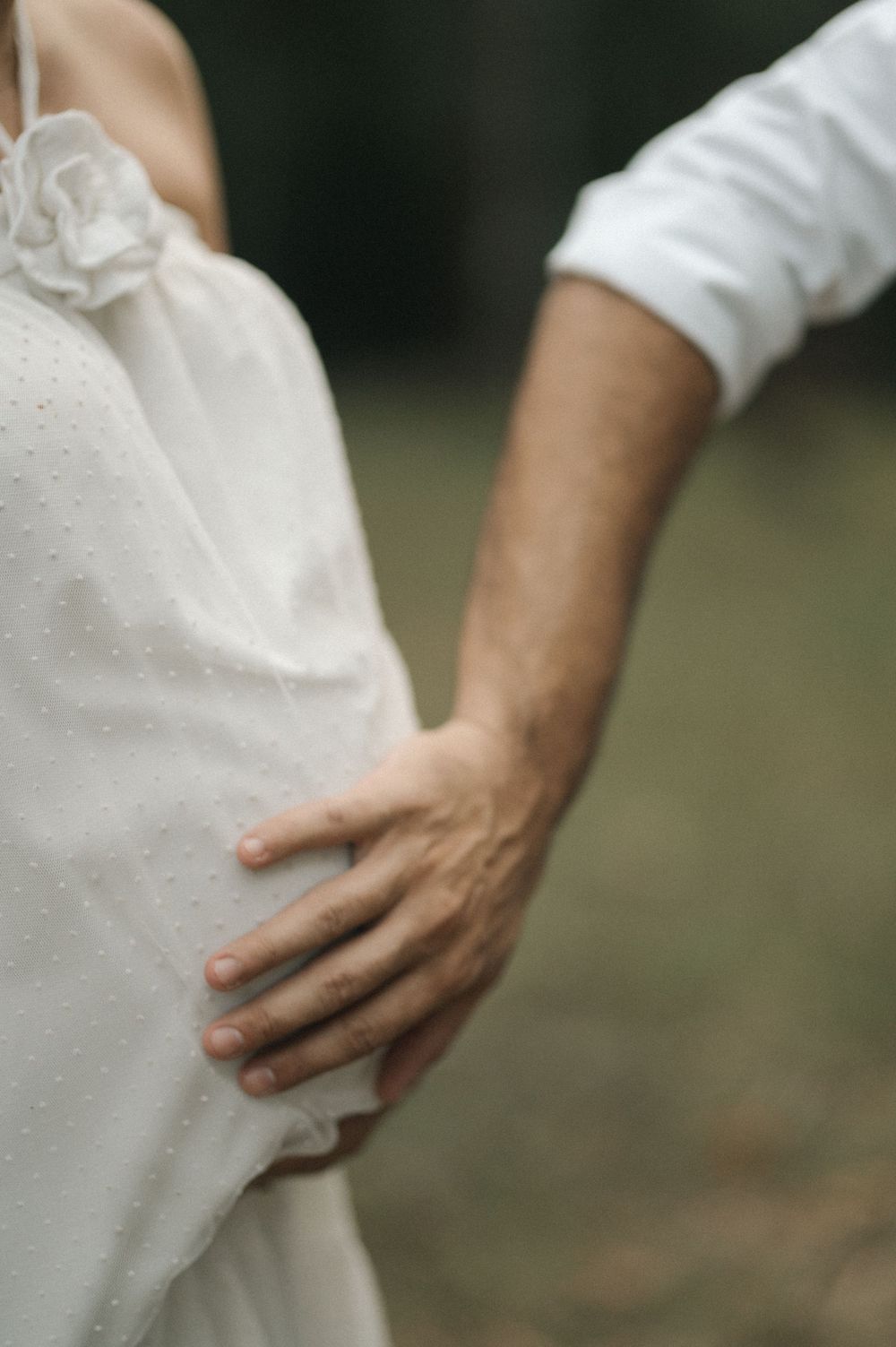 A gentle hand rests on white wedding dress fabric in a romantic outdoor setting.