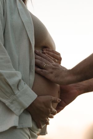 Close-up of hands tenderly cradling a pregnant belly against sunset background.
