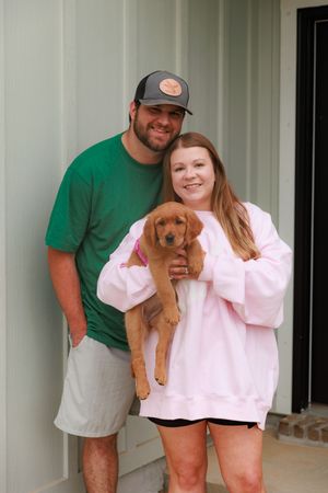 A couple poses with their small brown puppy on a porch with white columns.