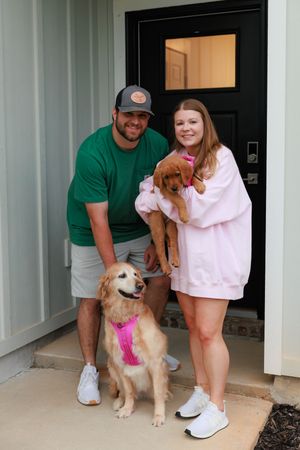 A couple stands with their two dogs, a small brown puppy and a golden retriever wearing a pink bandana, on their porch.