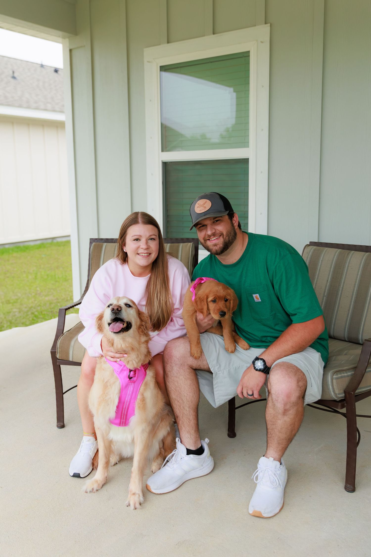 Family sits together with their golden retriever puppy on a porch bench outside their white-sided home.