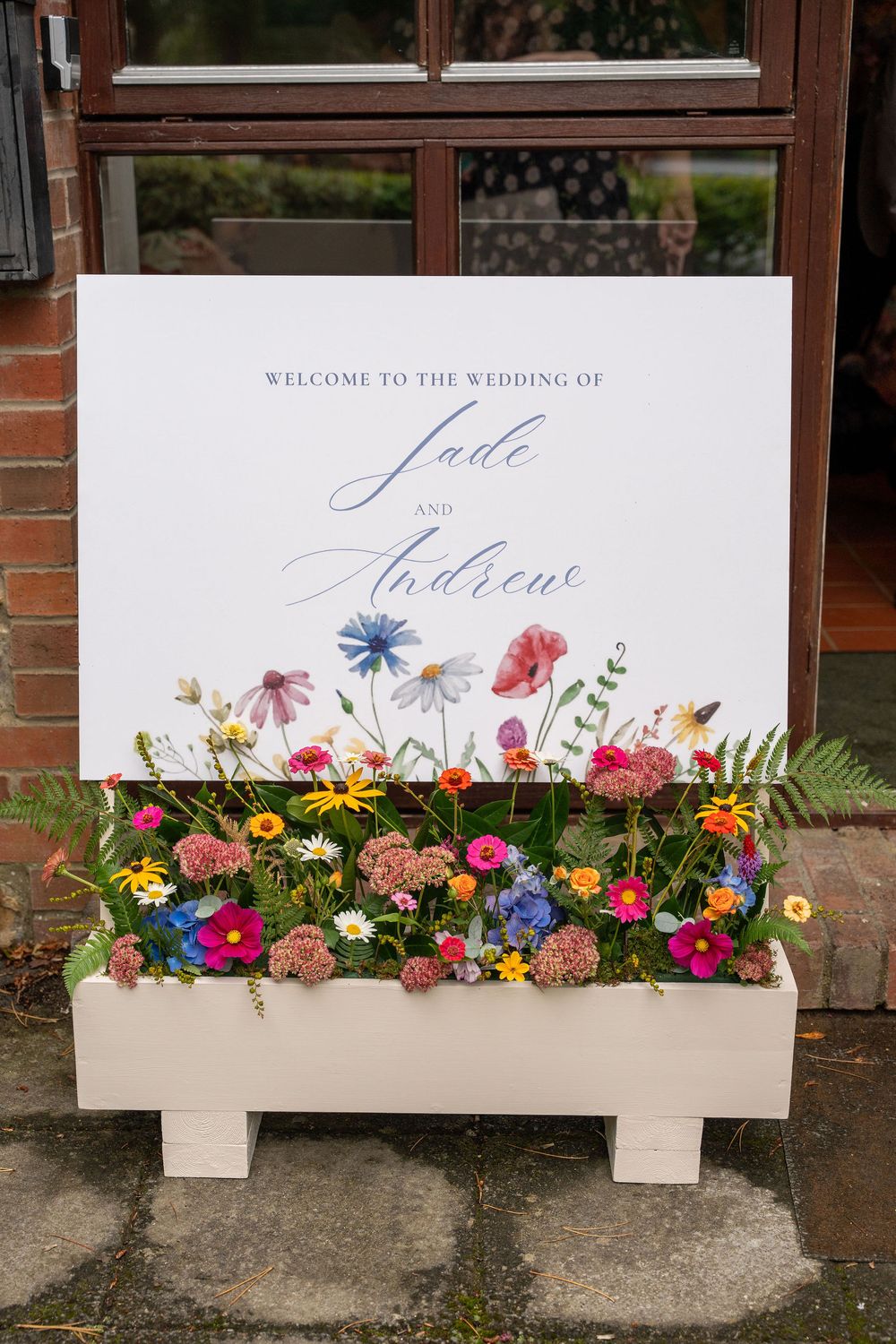 Wedding welcome sign with colorful wildflowers arranged along bottom edge against rustic brick wall backdrop.