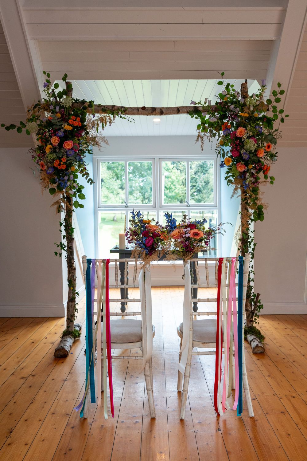 Floral wedding arch with colorful ribbons and flowers frames an elegant sweetheart table in a bright white room.