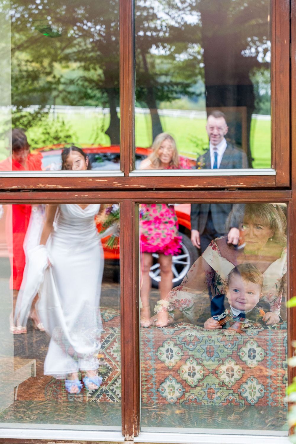 Wedding party guests visible through window panes of a rustic venue with a bride in white dress and colorful decorations.