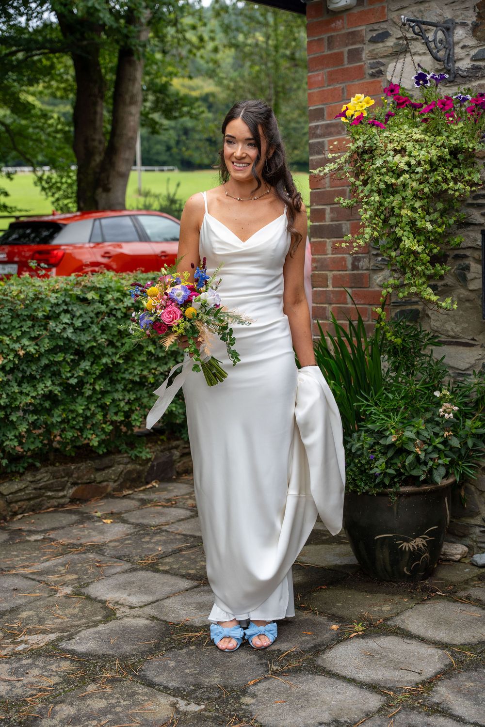 A person in a white wedding dress sits on stone steps holding a colorful bouquet of flowers near a brick wall.