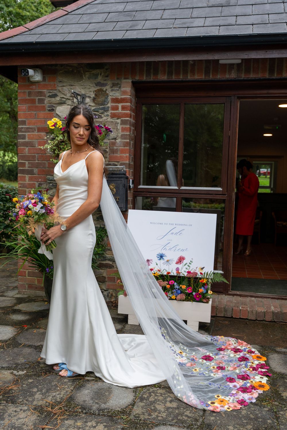A bride in a white wedding dress poses with a flowing veil decorated with colorful flower petals against a brick building.