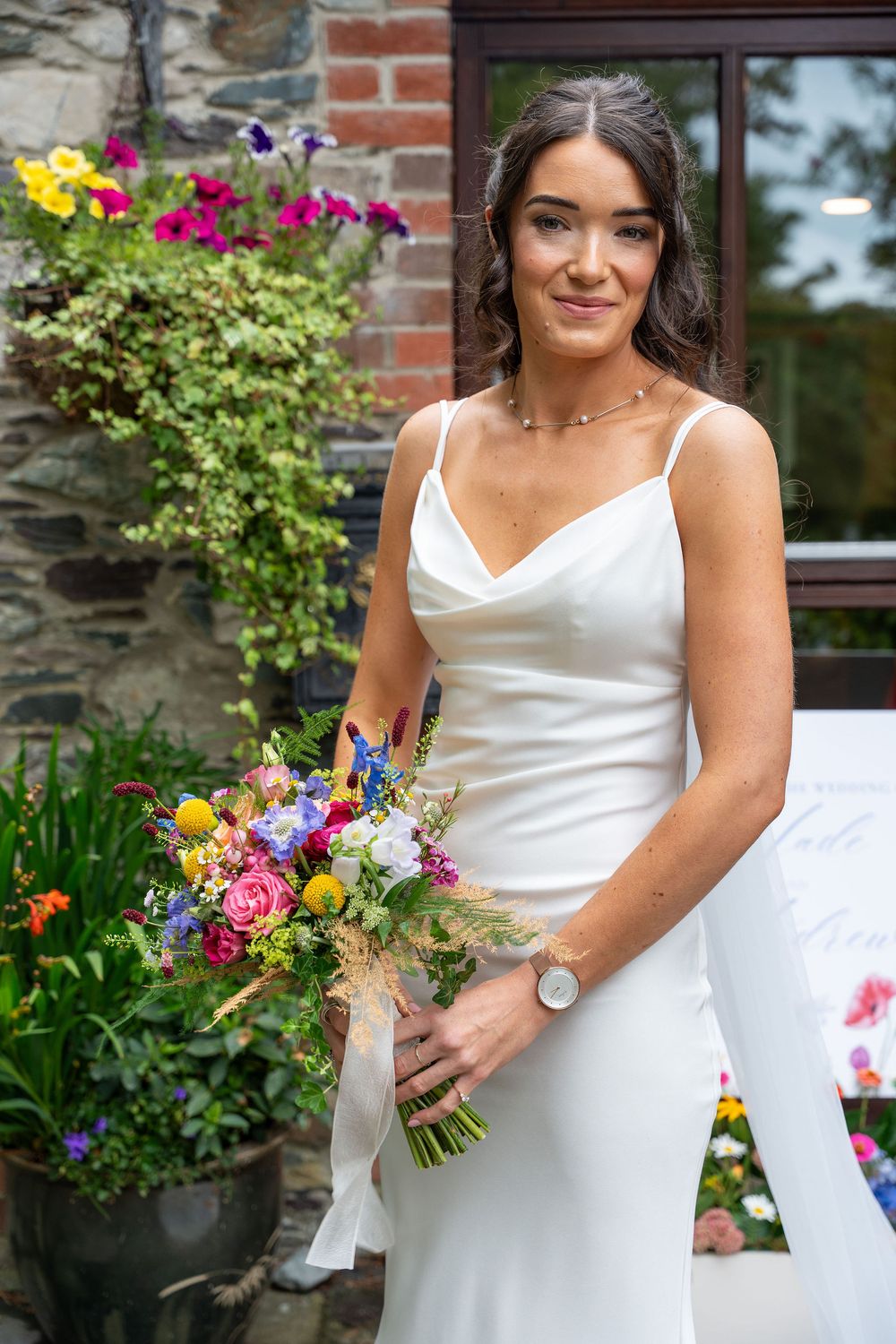 A bride in a white satin wedding dress holds a vibrant wildflower bouquet against a brick and garden backdrop.