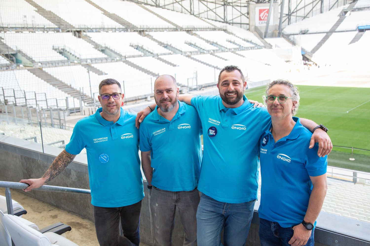 Four staff members wearing matching light blue polo shirts stand together in an empty stadium venue.
