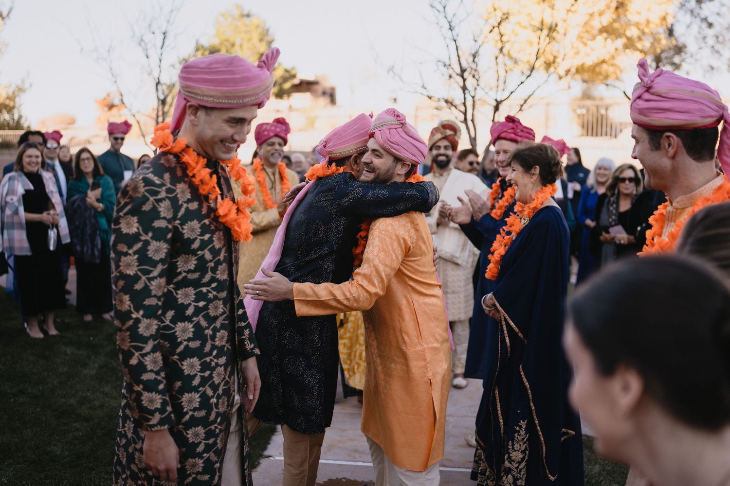 Men in colorful turbans and floral garlands embrace during a vibrant Indian wedding ceremony outdoors.at Sandia Resort and Casino