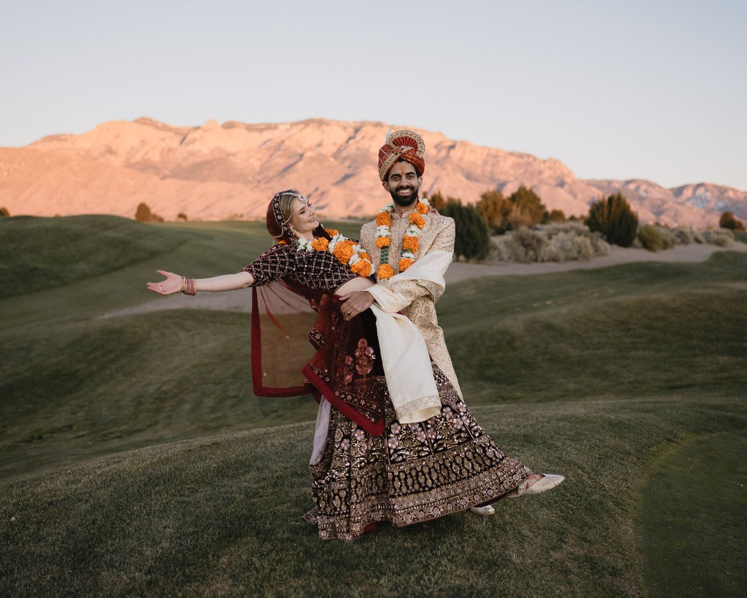 Indian wedding couple in traditional attire posing on green hillside with mountain backdrop at sunset.at Sandia Resort and Casino